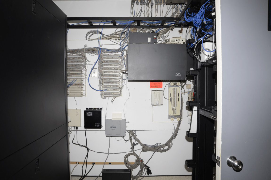 Une salle de serveurs encombrée est remplie de nombreux câbles, équipements et racks, suggérant une infrastructure complexe.